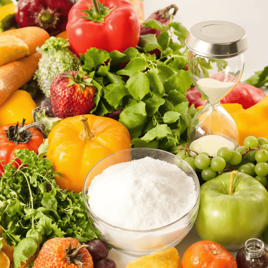 Close-up of fresh fruits and vegetables beside a sugar bowl with a cracked hourglass in the background, symbolizing sugar's impact on aging and health.