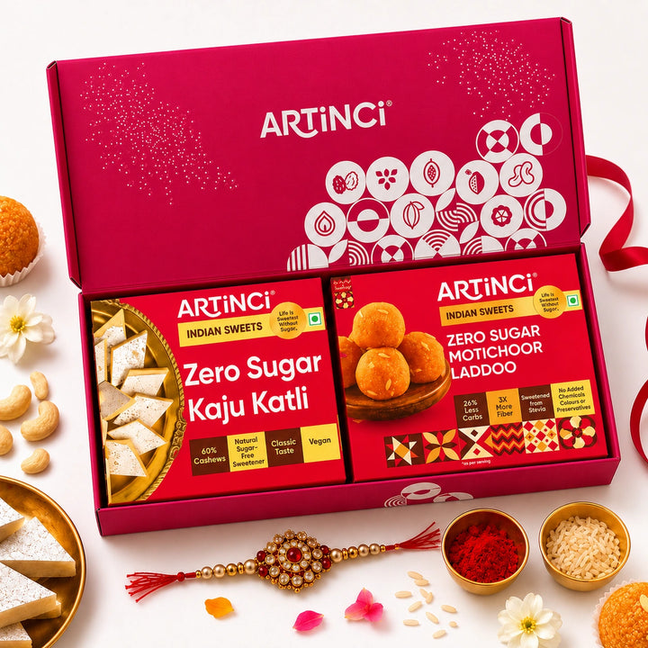 An open red ARTINCI gift box containing two packaged Indian sweets: Zero Sugar Kaju Katli and Zero Sugar Motichor Laddoo. The box is styled on a white surface with scattered cashews, flowers, a Rakhi, and small bowls of red powder and rice.