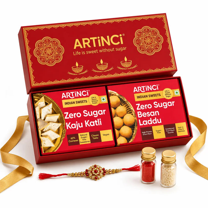 ArtiNci Indian Sweets gift set in a red box with gold detailing, shown open with Zero Sugar Kaju Katli and Zero Sugar Besan Laddu packets inside. A gold ribbon, a red and gold Rakhi, and two small spice jars are arranged in front on a white background.